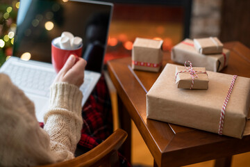 Christmas. Woman in sweater using laptop for searching gift ideas sitting at table near fireplace...