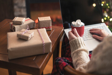 Fototapeta na wymiar Christmas. Woman in sweater using laptop for searching gift ideas sitting at table near fireplace and christmas tree with cup of cocoa and marshmallows. Concept