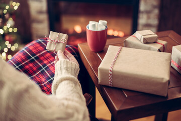 Fototapeta na wymiar Woman covered plaid sitting and relaxation near fareplace and christmas tree with cup of cocoa and marshmallows after finishing packing gift boxes for family.