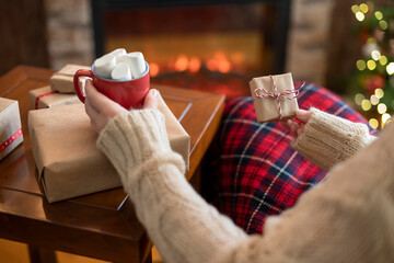 Fototapeta na wymiar Woman covered plaid sitting and relaxation near fareplace and christmas tree with cup of cocoa and marshmallows after finishing packing gift boxes for family.