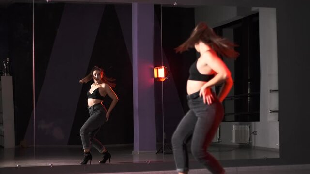 Young energetic woman in high heels dancing modern dance, performs plastic movements in a studio with large mirrors. 