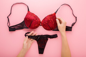 Young adult woman hands holding black red bra and string panties on light pink table background. Pastel color. Daily underwear. Closeup. Point of view shot. Top down view.