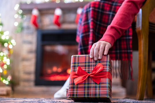 Woman legs in a winter socks covered plaid sitting and relaxation on armchair near fireplace and christmas tree pakking gift boxes for family. Bottom view.
