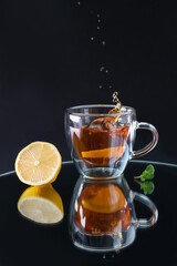 a glass cup with black tea, a splash in the cup from falling lemon, reflection on black background, lemon and mint leaf arranged on the table