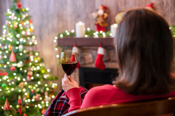 Back view of woman drinking wine from glass covered plaid sitting and relaxation on armchair near christmas fareplace and fir-tree.