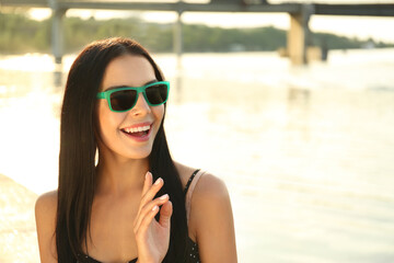 Beautiful young woman wearing stylish sunglasses near river. Space for text