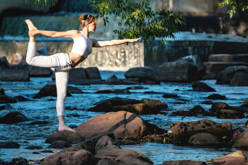 Young slender girl in white practicing yoga outdoors. Evening meditation and relaxation at the river and waterfall. The Dancer Pose of yoga (Natarajasana).