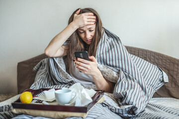 Young sick woman is sitting in bed wrapped in a blanket, holding her aching head and using her phone