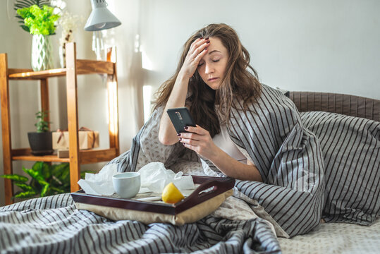 Young sick woman is sitting in bed wrapped in a blanket, holding her aching head and using her phone