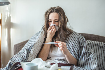 Young sick woman is sitting in bed wrapped in a blanket and using a handkerchief and looking at the...