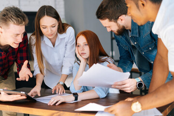 Side view of exhausted tired young business woman sitting at desk, colleagues shaking papers in face paper documents. Businesswoman under stress at workplace. Female boss exhausted by management work.