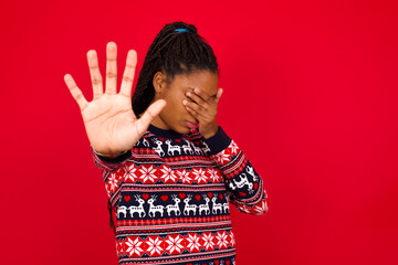 Young beautiful African American woman standing against red background covers eyes with palm and doing stop gesture, tries to hide from everybody.