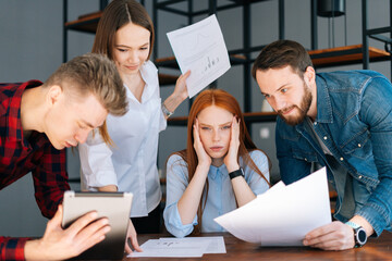 Portrait of tired business woman sitting at table, looking at camera, multi-ethnic colleagues shaking paper documents. Multiethnic business team wants to get answer and decision from boss.