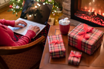 Christmas. Woman using laptop for searching gift ideas sitting at table with ready gift boxes and cup of cocoa and marshmallows near fireplace and christmas tree. Concept