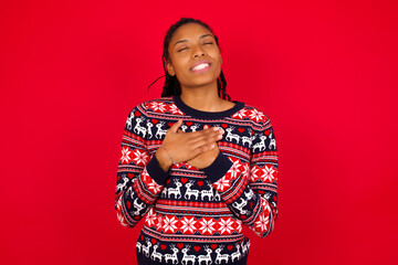 Young beautiful African American woman standing against red background smiling with hands on chest with closed eyes and grateful gesture on face. Health concept.