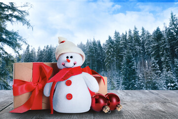 Cute toy snowman with gift boxes and Christmas balls on wooden table in winter forest. Space for...