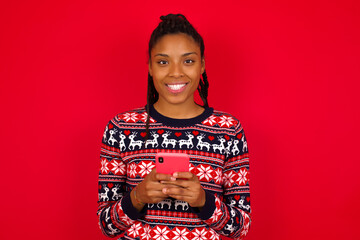 Young beautiful African American woman standing against red background enjoys distant communication, uses mobile phone, surfs fast unlimited internet, has pleasant smile, makes shopping online,