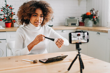 Dark skinned blogger holding professional makeup brush and showing tone on hand sitting against smartphone on tripod closeup