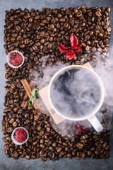 White cup full of coffee beans on a coffee beans background with red chocolate candy and red flowers. Morning espresso. Coffee mug.	