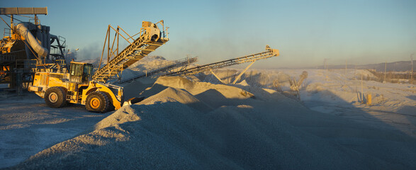Front-end loader on a background of stone crushing equipment in the mining enterprise in the rays of the setting sun, panorama. Industrial landscape.