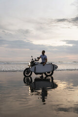 Surfer rides on motorbike with surfboard at sunset ocean beach. Bali island, Indonesia