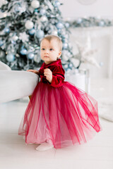 Cute little baby girl in red dress standing near the chair on the background of beautiful Christmas tree and decorations. Christmas, family, New Year concept
