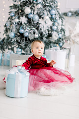 Cute happy little girl in wine red dress plays with Christmas box gift and sits on the floor near a decorated Christmas tree. Family, Christmas, New year concept