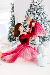 Family Christmas at cozy decorated interior. Young mother in red dress sits near the Christmas tree and holds her cute little daughter in her arms. New year concept.