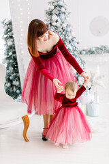 The first steps of cute little beautiful girl, with pretty joyful elegant mother, supporting her, having fun while celebrating winter holidays at light Christmas room interior