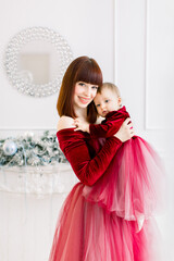 Christmas and Happy New Year Holidays. Family, mother and child concept. Tender photo of happy pretty mom holding and hugging her cute baby girl in red dress, having fun at decorated room