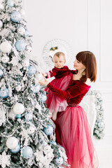 Charming young Caucasian mother holding her cute little daughter and decorating christmas tree at beautiful light decorated studio. New Year and Christmas concept