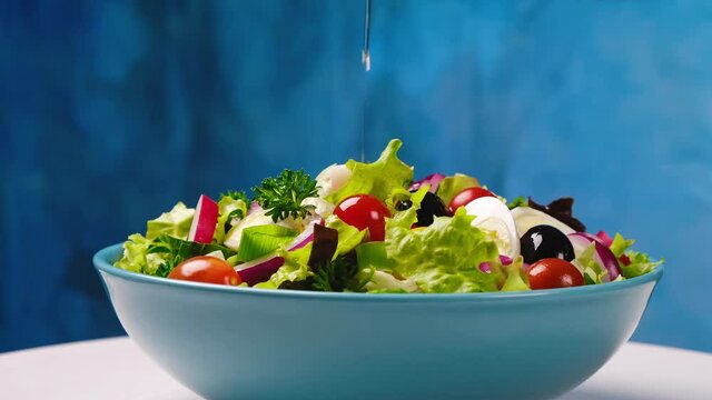 Olive oil pouring from bottle onto vegetable salad, healthy and dietary food from fresh ingredients in bowl on blue background, slow motion	
