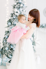 Portrait of smiling young mother holding her beautiful little baby girl, posing on the background of decorated Christmas tree. Family time, winter holidays concept