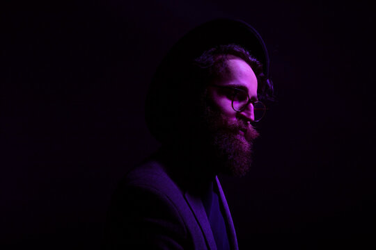 Portrait of a bearded man in hat and eyeglasses posing in profile in studio, isolated on dark neon purple background.