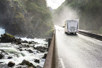 Motorhome Camper Van driving fast on wet canyon valley road and bridge across waterfall river. Norway.