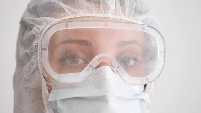 Close up of young female doctor wearing full PPE protective gear against coronavirus covid-19 virus, looks directly into c amera and blinks. Slow motion with white background.