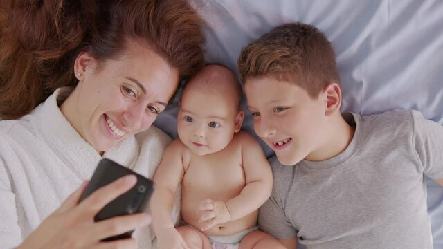 Happy mother with her kids are making a selfie or video call to father or relatives in a bed. Concept of technology, new generation,family, connection, parenthood, authenticity.