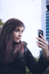 Young latin brunette woman dressed with dark clothes, uses a mobile phone to take selfies, showing emotions. Photo taken in the financial district, surrounded with skyscrapers during the winter season