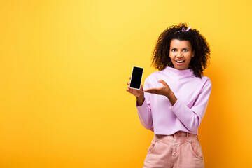 Naklejka premium excited african american woman pointing with hand at smartphone with blank screen on yellow