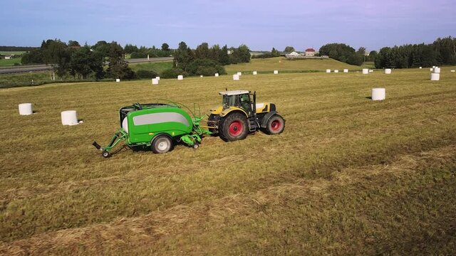 Tractor with trailed bale machine works on the field. Agricultural work, baling, baler, hay collection in the summer field. Parallel panoramic movement 2.7K drone footage