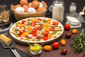 Pizza with ingredients is ready to bake. Cheese grater on table. Tomatoes and sprig of rosemary. Egg basket, jar and flour sieve
