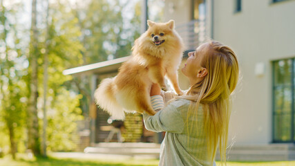 Young Beautiful Woman Plays with Her Cute Little Pomeranian Dog, Holds Her, Caresses, Cuddles, and Almost Kisses. Gorgeous Sunny day, Having Fun on the Idyllic House Lawn