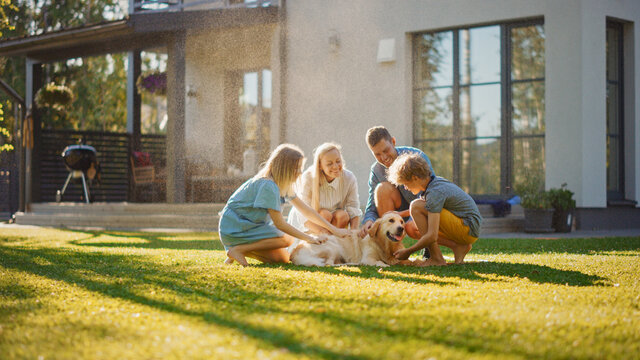 Beautiful Smiling Family Of Four Cuddling Happy Golden Retriever Dog On The Backyard Lawn. Idyllic Family Cuddling Loyal Pedigree Dog Outdoors In Summer House Backyard.