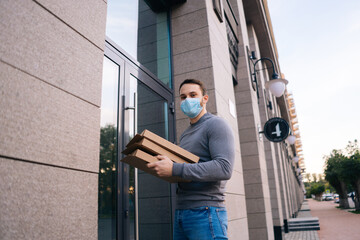 Low-angle shot of delivery man wearing medical mask ringing door for delivery carton boxes with hot pizza. Deliveryman in protective mask holding box with food. Concept of delivery during quarantine.