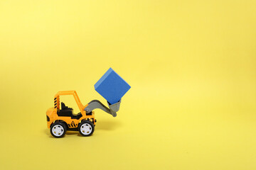A toy bulldozer lifting a cube on yellow background 