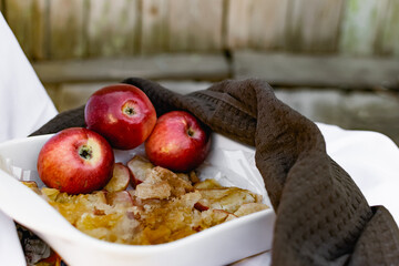 Collapsed soft baked apples in a white ceramic square shape on a special package. A white tablecloth is laid carelessly on the table, next to a dark napkin and ripe juicy red apples