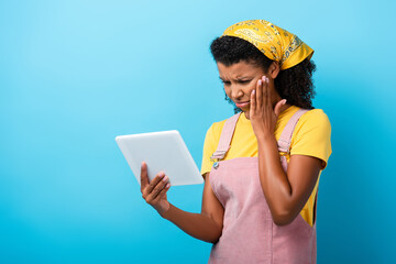 upset african american woman in headscarf using digital tablet on blue