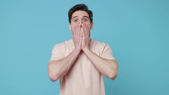 Shocked surprised young man 20s years old in casual beige t-shirt posing isolated on pastel blue colour background in studio. People lifestyle concept. Looking camera say wow covering mouth with hands
