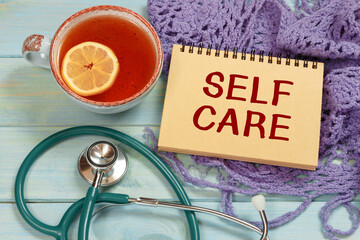 Notepad with text SELF CARE on a table. Hot tea, knitted scarf and stethoscope.
