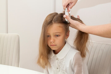 Close up view of calm sad schoolgirl schoolgirl with long hair and her mother who is combing her hair at home, they are dressed in white blouses, lifestyle people concept